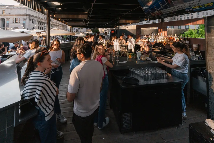 The rooftop at golden hour, a crowd three deep at the bar with Moynihan Train Hall behind them